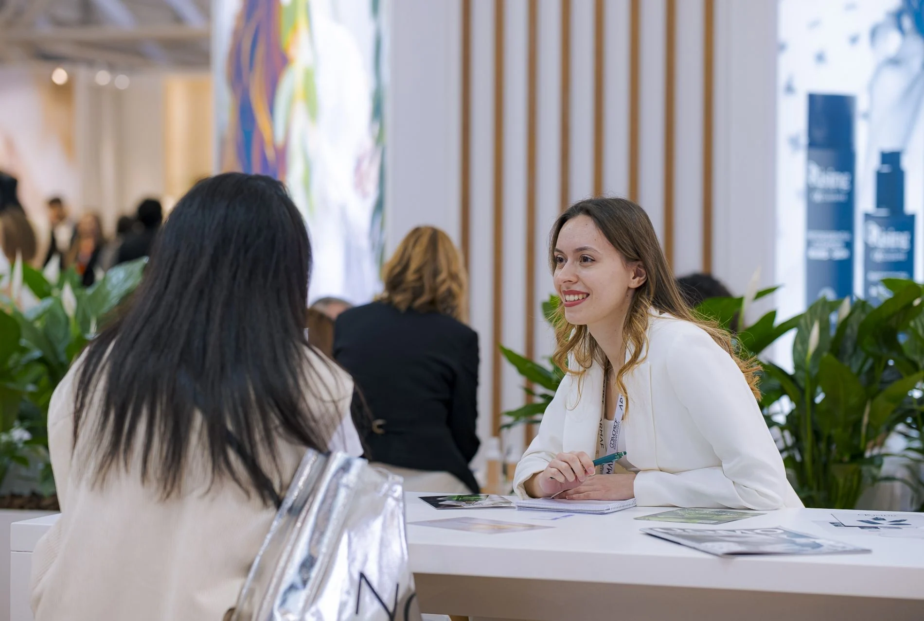 Beauty professionals holding a business conversation at a plant-filled exhibition stand.