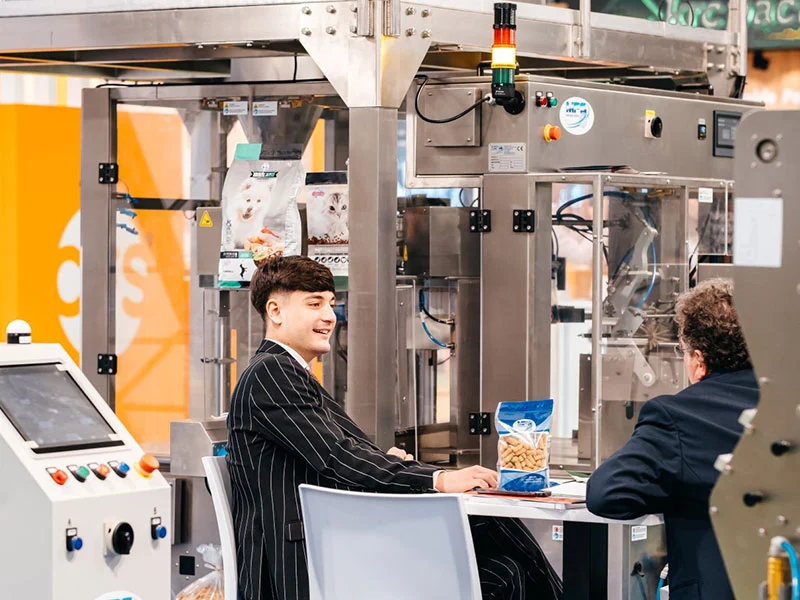 Two professionals meet beside operating pet-product packaging machinery in the Zoomark Factory area.